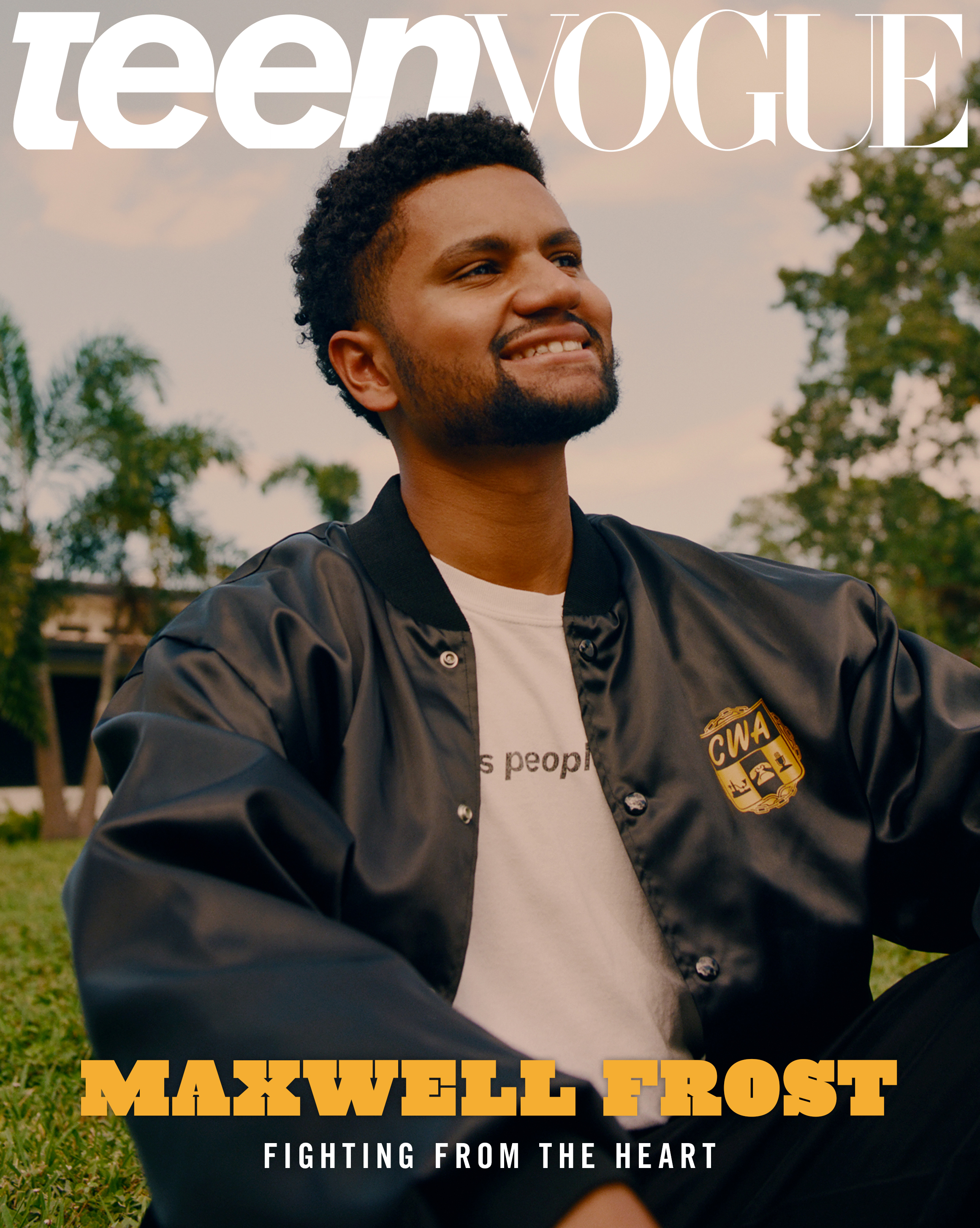 Congressman Maxwell Frost on the cover of Teen Vogue. He's sitting on a lawn while smiling and wearing a white t-shirt and black bomber jacket. 