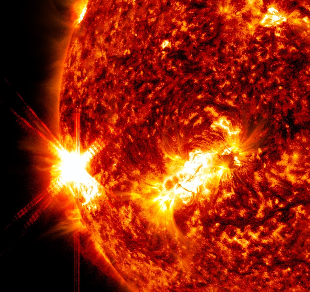 A red, gold, yellow, white, and black Sun on a black background. On the left, there is a white and yellow "X" coming off of the Sun. That is the solar flare. To the right of the X is a splotch of white and yellow. The rest of the Sun is a mix of black and red.