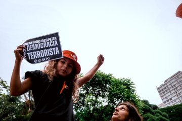 Uma criança está com uma camiseta preta com um detalhe de um arco-íris, um chapéu de pescador escrito Lula, segurando uma folha em que está escrito "quem não respeita a democracia é terrorista" enquanto a outra mão está levantada com o punho cerrado.