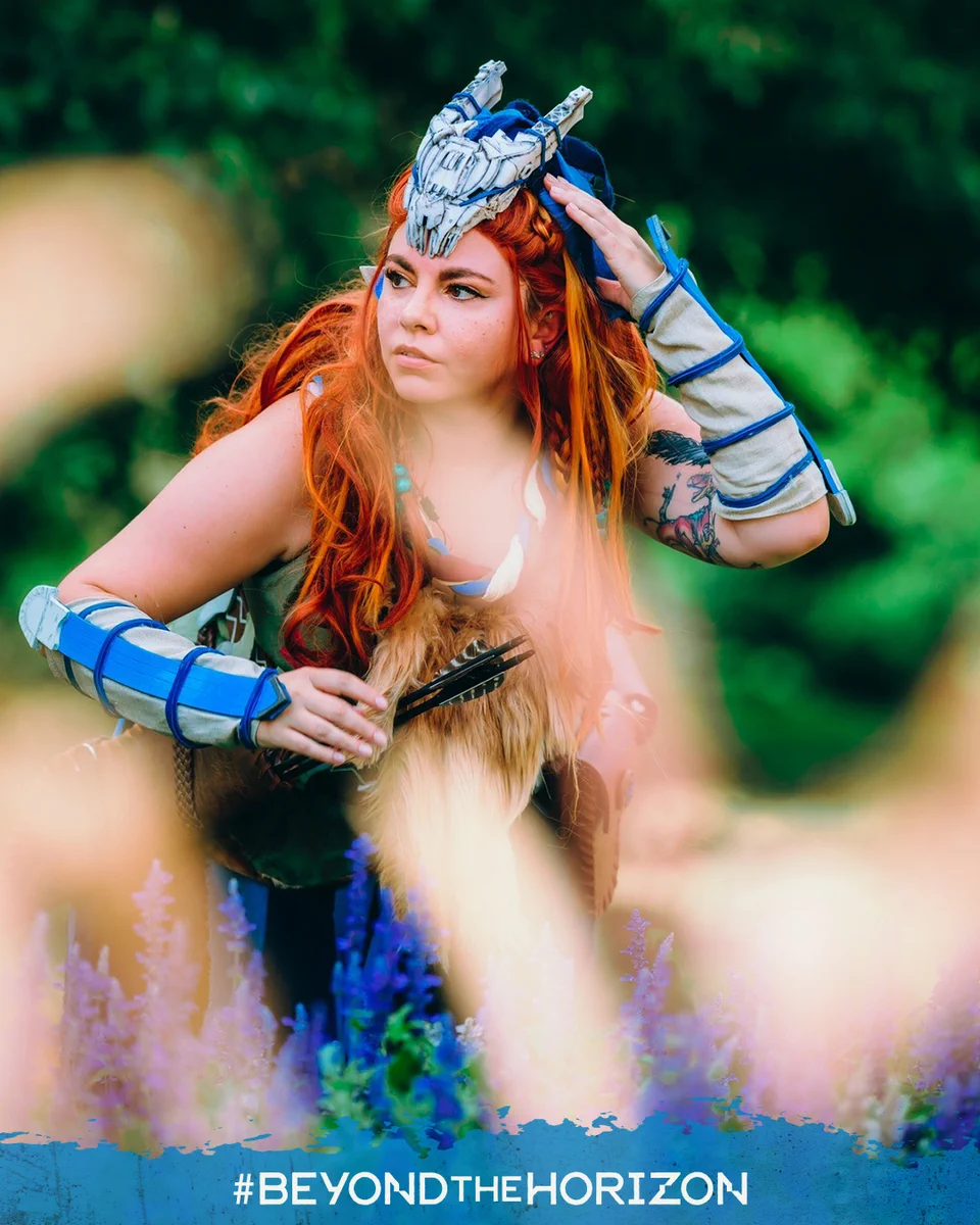 A cosplayer is dressed as Aloy from the Horizon franchise. Dressed in the Banuk Sickness Eater armor, this armor is designed to protect Aloy against plummeting temperatures and Corruption damage. However, this cosplayer is an a lush green setting, with blue and purple flowers sticking up around her. This allows her vibrant red hair to stand out in the photo.