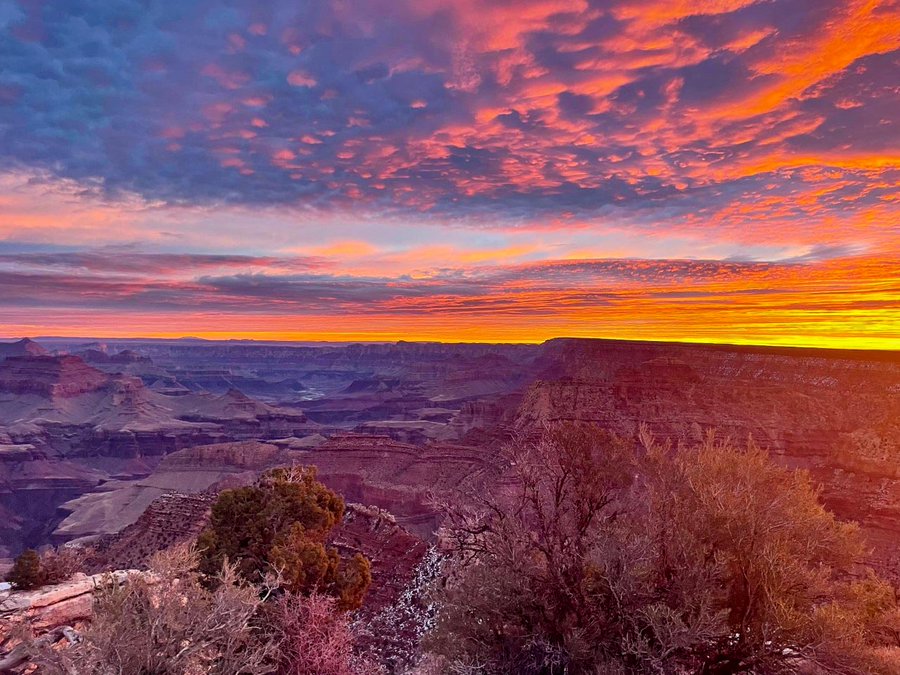 patchy clouds in the sky are tinted shades of brilliant orange and yellow at sunrise above a vast canyon landscape filled with cliffs, peaks and ridgelines. 