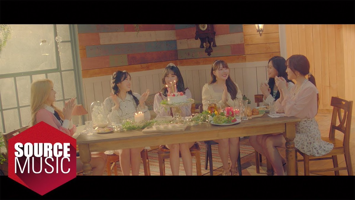 A group of young women celebrate a birthday around a candlelit cake at a wooden table in a rustic room; the image features the red "SOURCE MUSIC" logo in the bottom left corner.