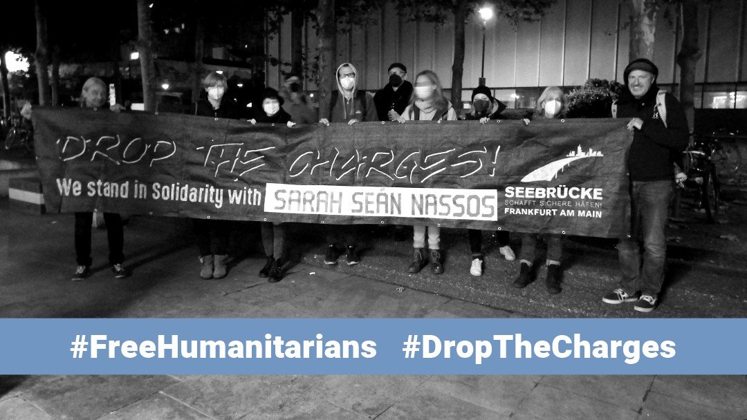 SW Foto von Aktivisti mit Transpi:<br>DROP THE CHARGES<br>We stand in Solidarity with SARAH, SEÁN & NASSOS<br>u. Seebrücke FFM Logo