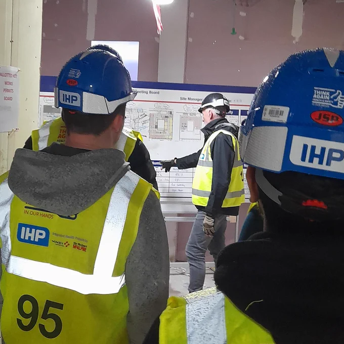Learners in hi-vis jackets and helmets watch a Construction worker, who is pointing to a diagram on a board.