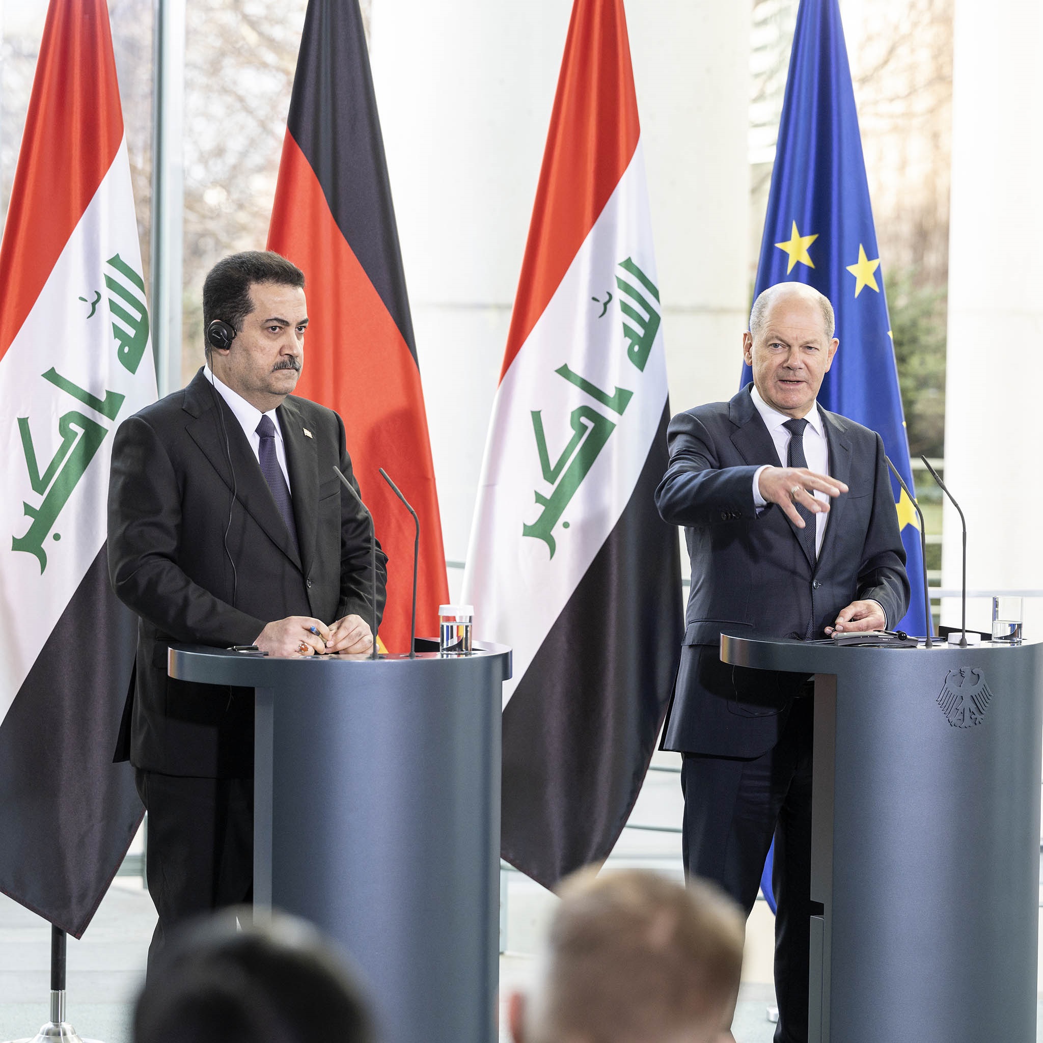 Bundeskanzler Olaf Scholz gibt gemeinsam mit dem irakischen Premierminister Sudani eine Pressekonferenz im Bundeskanzleramt. Das Foto zeigt beide nebeneinander jeweils am Pult stehend. Bundeskanzler Olaf Scholz beantwortet gerade eine Frage und gestikuliert dabei mit der Hand. Im Hintergrund sind die Staatsflaggen Deutschlands und Iraks sichtbar. 