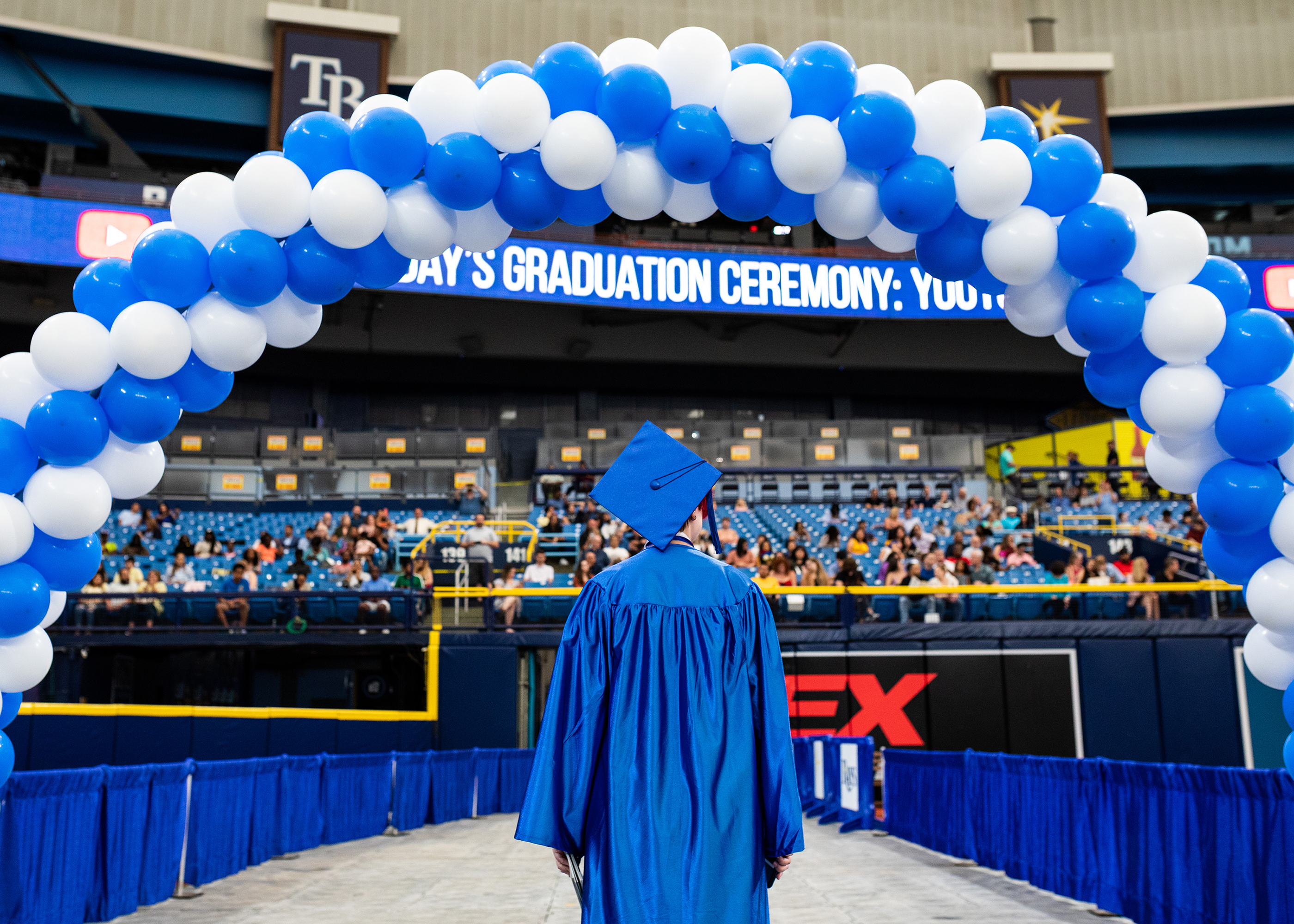 A student in a blue graduation cap and gown standing under an archway of blue and white balloons.