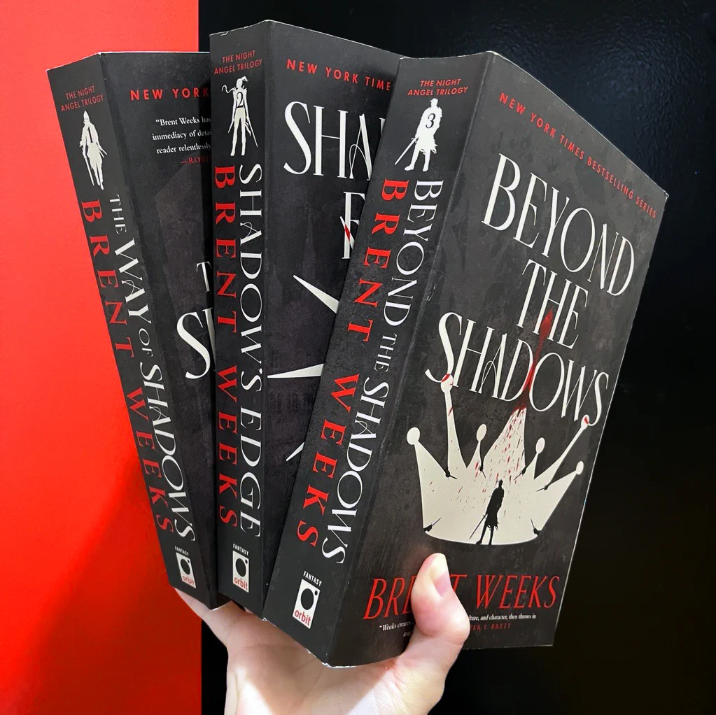 A hand holding The Way of Shadows TP, Shadow's Edge TP, and Beyond the Shadows TP by Brent Weeks against a black door with a red stripe