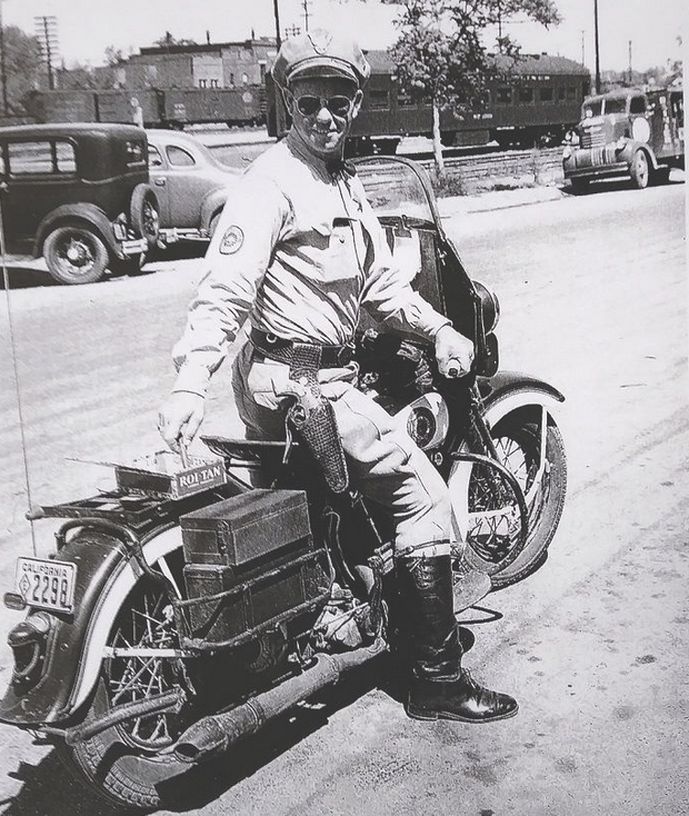 A CHP motorcycle cop pulls a cigar from the back of his bike, c. 1950. He had an ashtray there as well. This is in Roseville on Vernon Street when it was Highway 40, the transcontinental Highway. In the background are the Roseville Rail Yards.