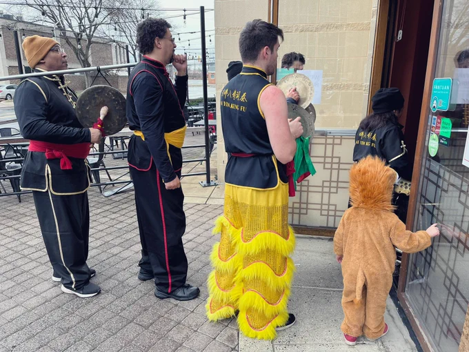 Chinese lion dance drummers in their black Kung Fu outfits and playing cymbals stand in the doorway of a Chinese restaurant, clearly playing for lion dancers which aren’t visible isn’t the restaurant. Amongst them stands the 3-year-old in her lion costume, peering in through the doorway.