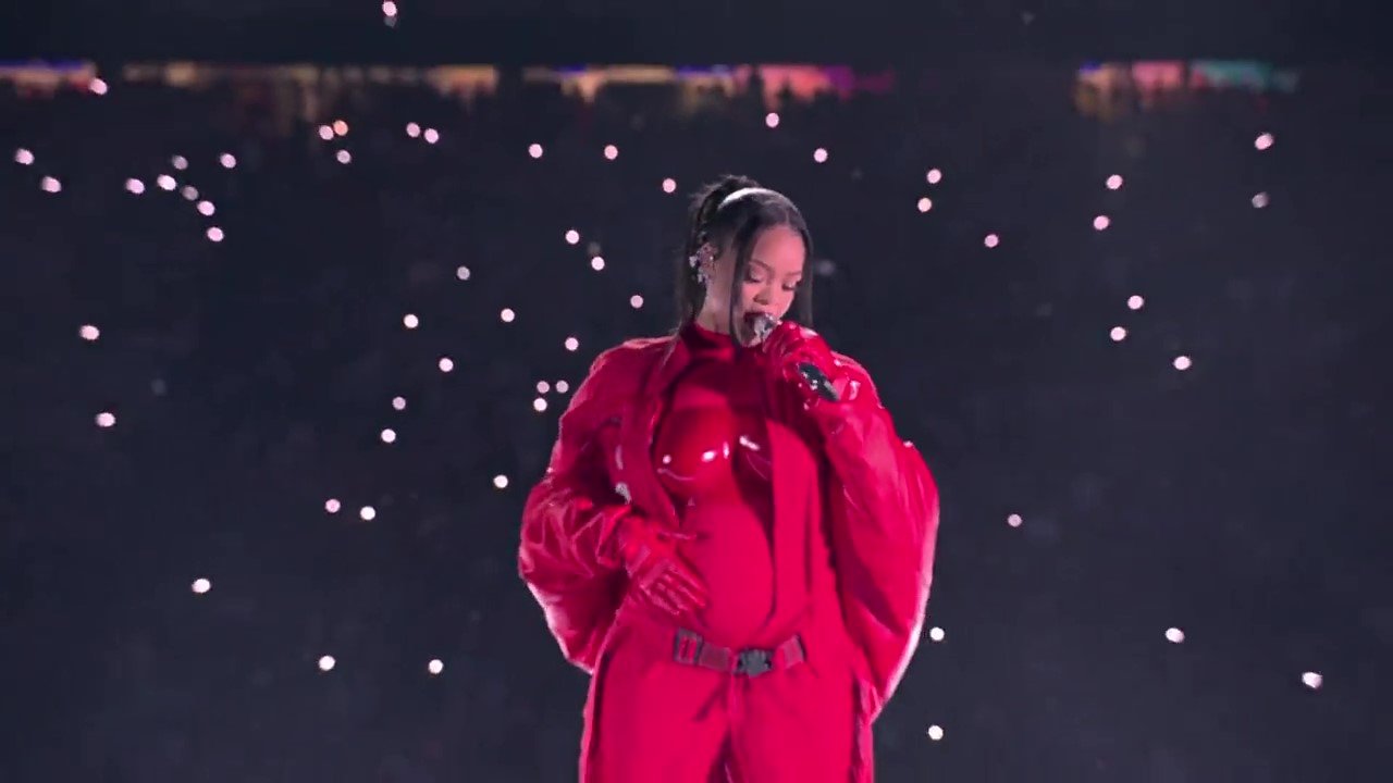 Rihanna singing onstage at the Super Bowl, resting her arm on her stomach.