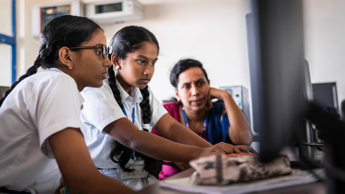 Photo of two students and a teacher sitting at a computer looking at the screen.