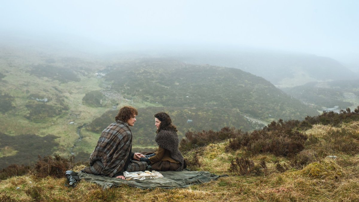 Outlander's Jamie and Claire sit on a rug on the top of a hill in the Scottish highlands.