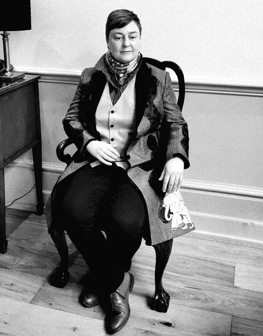 A black and white photograph of Rachel Dax. A woman with a short, brown pixie cut is sat in a wooden chair. She's wearing a neck-scarf, a waistcoat, and a long, embroidered jacket.