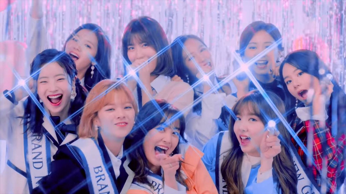 A K-pop group of young women celebrating with bright stage lights creating star-burst effects, smiling and holding microphones, wearing sashes that read 'GRAND' and 'BRAND'.