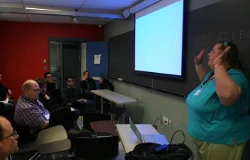 A group of people gathered in a classroom, facing a projector screen. At far right, you can see a woman talking with her arms in the air.