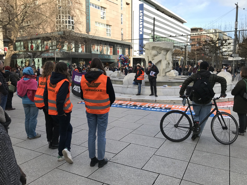 Menschen hören einem Redner am Brockhausbrunnen auf der Zeil in Frankfurt zu. Einige tragen orange Westen mit der Aufschrift 'Seebrücke'. Um den Brunnen Transpis.