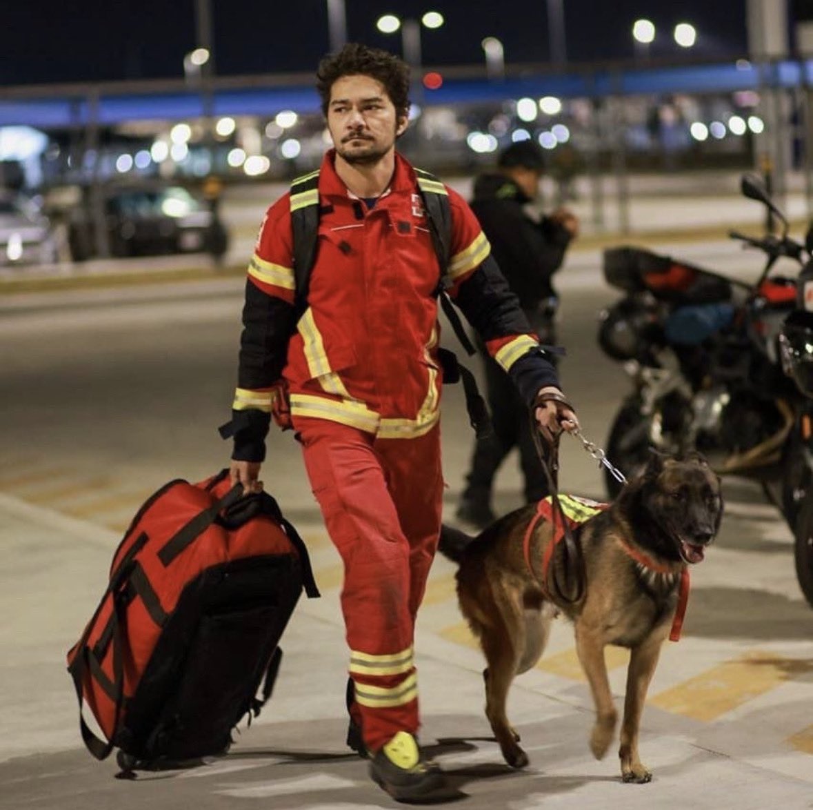 a member of the Red Cross wears a reflective, red uniform pulls a large red and black bag with his right hand, and the leash of a belgian malinois in his left hand. the dog walks with his handler across a parking lot.