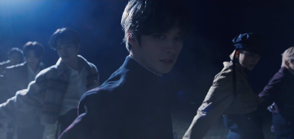 A scene from a K-pop music video featuring several male performers, with one in focus in the foreground under blue lighting.