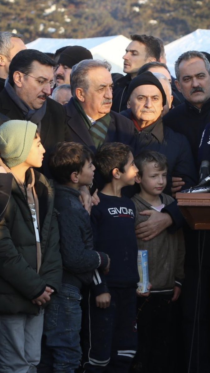 Paltolu atkılı bereli devlet adamlarının önünde montsuz çocuklar.