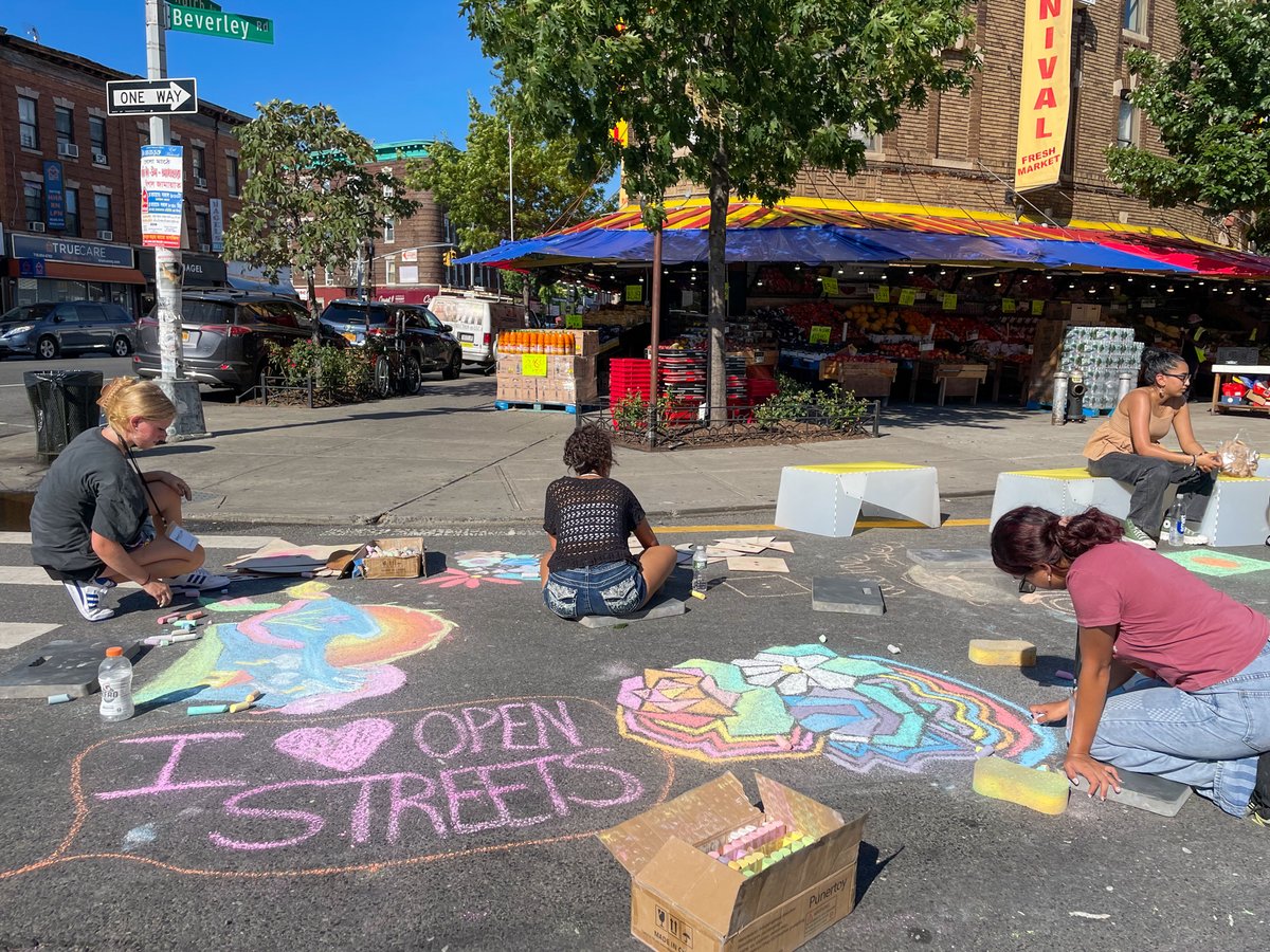 People use chalk to draw on a roadway that has been closed to traffic. The colorful drawings are abstract and one says I heart open streets
