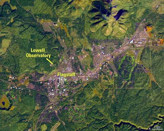 green hills and mountains border the frame of this satellite image of flagstaff. In the center, Flagstaff Arizona is brownish-gray with white roofs occasionaly popping through.
