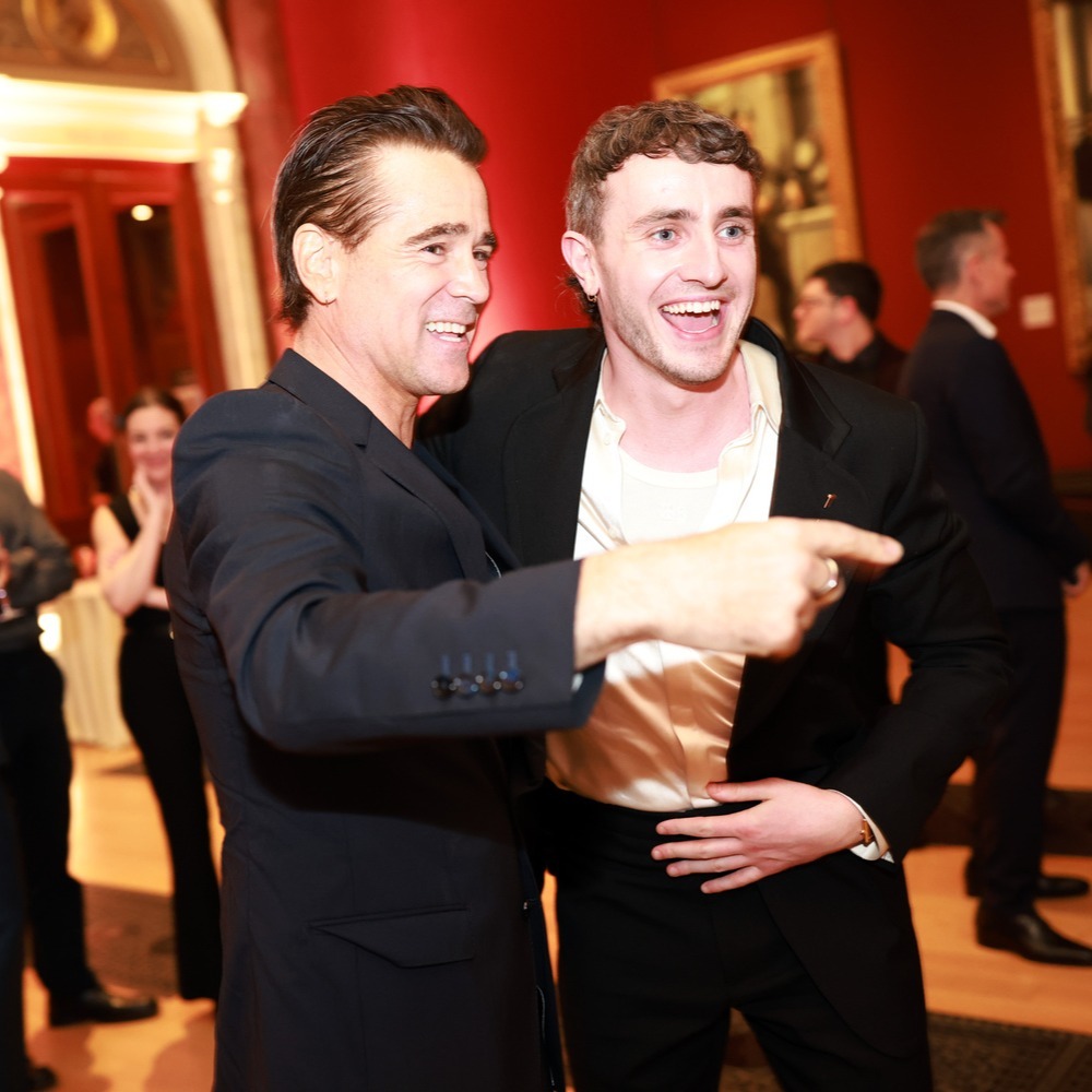 A shallow focus photo of actors Colin Farrell and Paul Mescal at the BAFTA Nominees party in London. They are both very happy to be there, hands on each other's backs. Colin is pointing to someone out of the frame. Paul has a big grin on his face. They are both looking smart in black suit jackets. In the background you can see the red walls of the National Gallery, some paintings and assorted attendees.