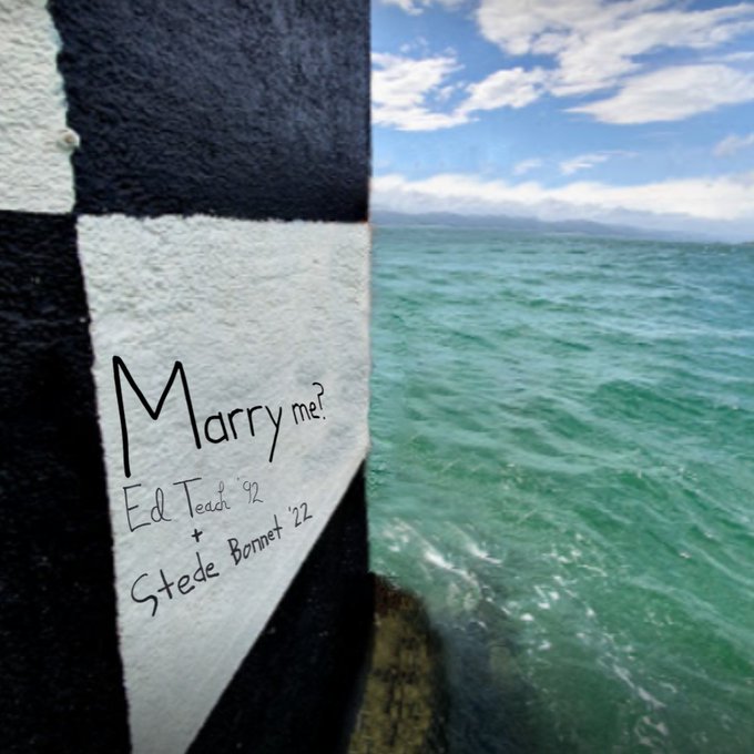 A close up photo of the black and white checked side of the lighthouse. The water is blue green. On a white square of the structure, "Ed Teach '92 + Stede Bonnet' 22" is written. Above that, in larger font, it says "Marry me?" 