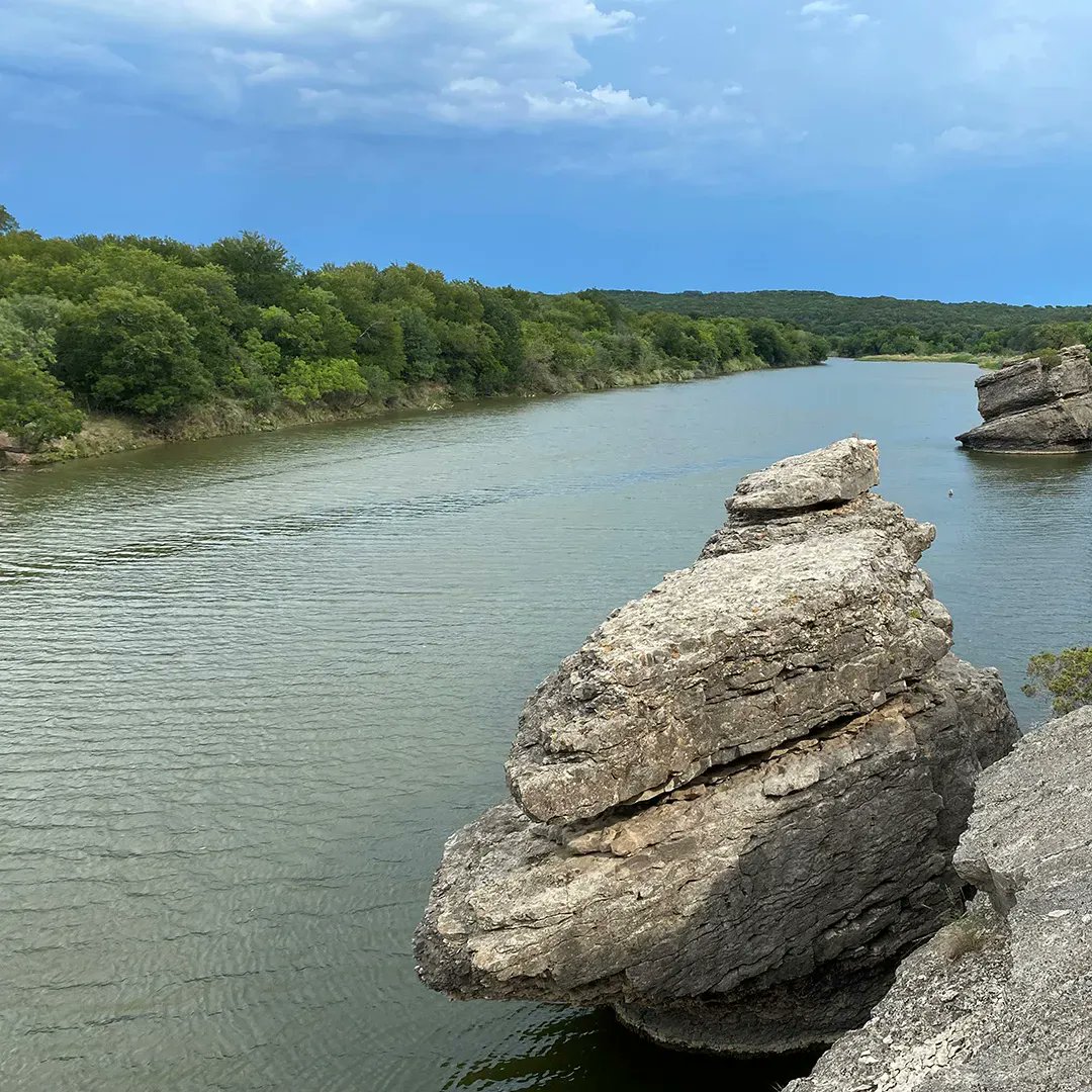 Image of the Brazos River with a large boulder in the foreground and trees and shoreline in the background.