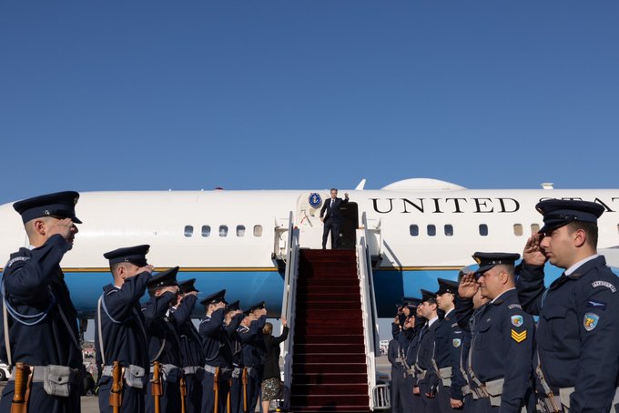 Secretary Antony Blinken waves on the top of the stairs as he boards the U.S. official plane. U.S. Military troop members in uniform stand on both sides of the stairs holding a salute. 