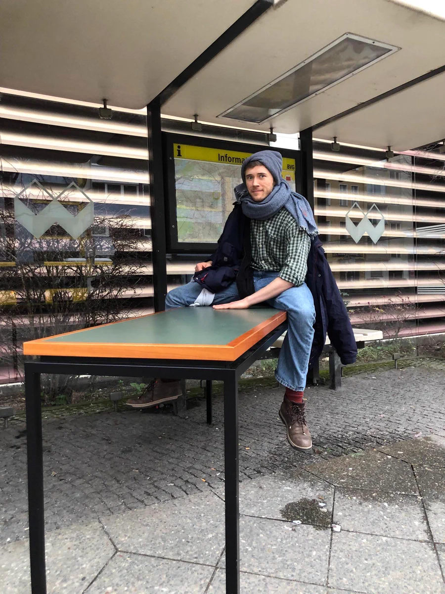 Ein langer Tisch mit schwarzen Beinen und einer grün-braunen Platte steht an einer Bushaltestelle. Henning sitzt rittlings darauf, die linke Hand liegt auf der Platte auf, er guckt in die Kamera. 