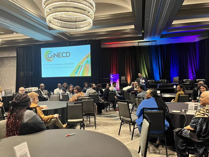 Ballroom with several people seated at chairs. In background is the CoNECD logo on a slide.