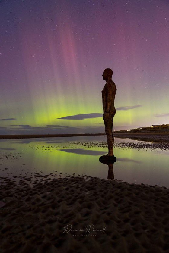 An Antony Gormley's 'Another Place' sculpture stands in the foreground of the picture on Crosby Beach in Liverpool.  The sculpture looks out on the scene, as he stands in a shallow tide that reflects the beautiful view above. The sky is a melody of colour, with purples, pinks, and yellows forming beautiful lines through the sky. Stars are scattered in the distance.