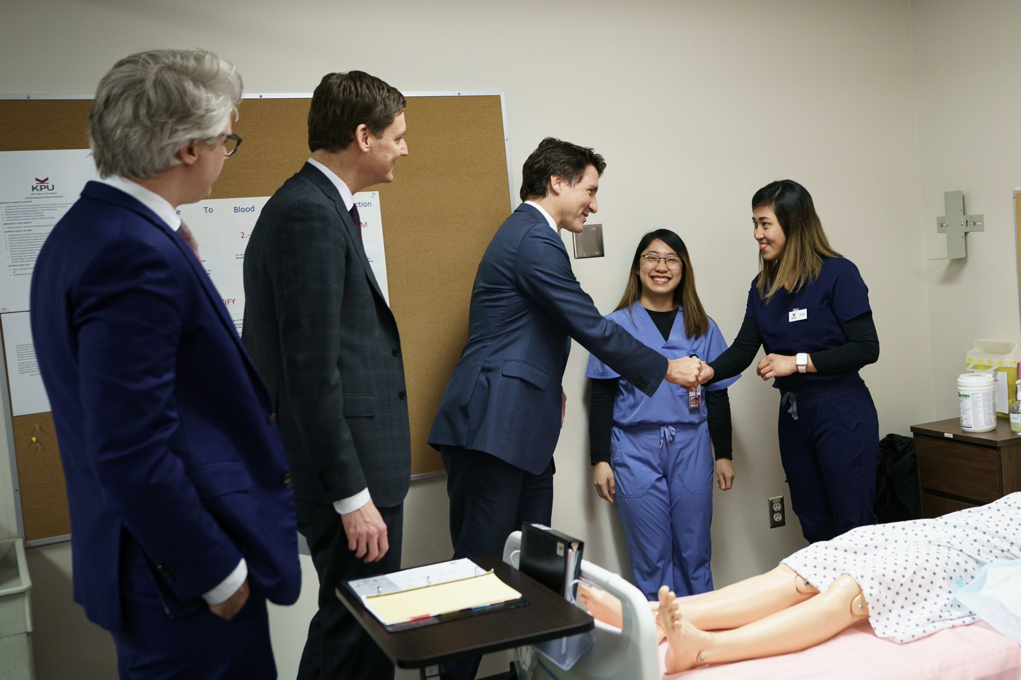 Prime Minister Justin Trudeau is standing inside a classroom and shaking hands with a nursing student. Premier David Eby, Minister Andrew Mercier, and another nursing student are standing beside them. On a bed in the foreground, a medical mannequin is laying down.