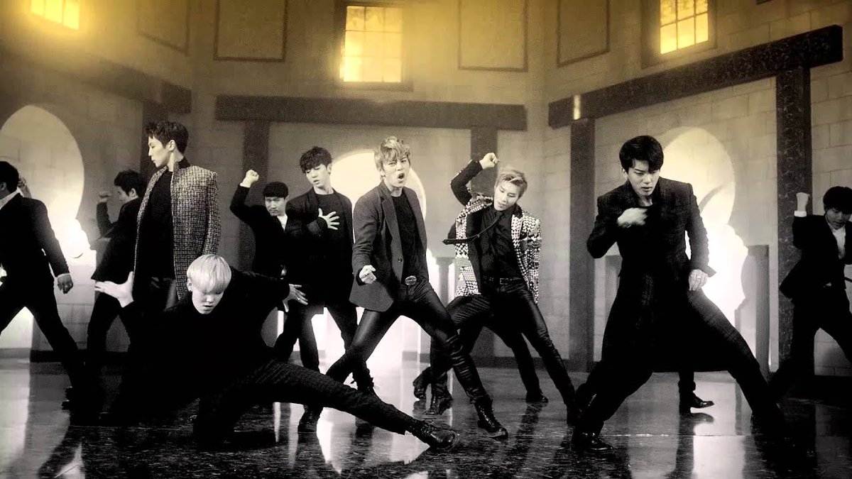 A group of men in suits perform a choreographed dance routine in a dimly lit, castle-like room with arched windows.