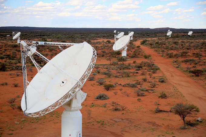 A large, white dish-like antenna in the foreground, and several more antennas in the background, sitting on red Earth surrounded by low vegetation; above is a blue sky with white clouds.