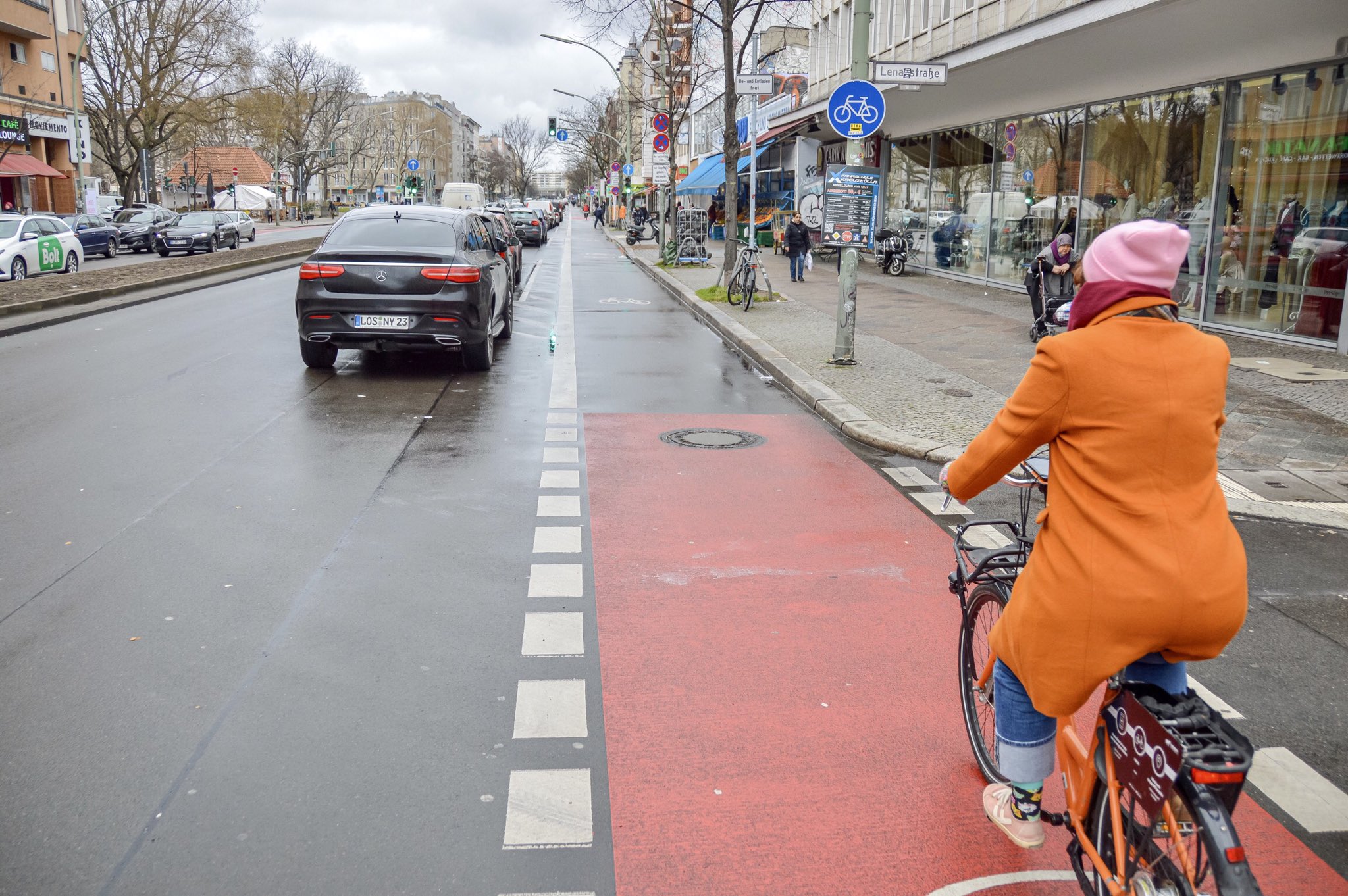 A female cyclist in casual winter attire rides in a protected bike lane lined with parked cars on one side and retail shops on the other in Berlin.
