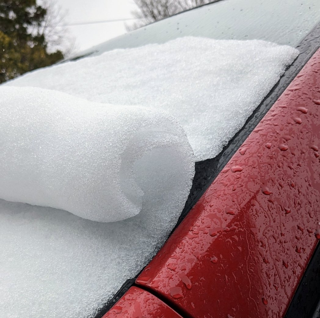 A thin sheet of snow curls like a wave on top of a car windshield