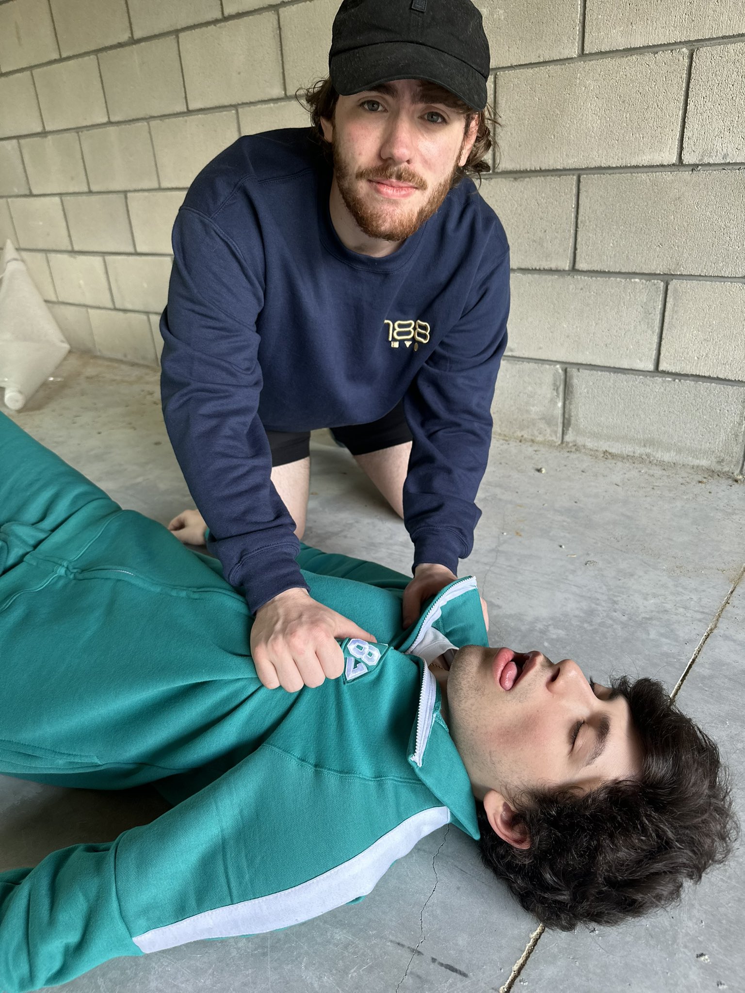 Sapnap holding GeorgeNotFound to the ground while he is acting dead. George is wearing Squid Games outfit and Sapnap is wearing his merchandise.