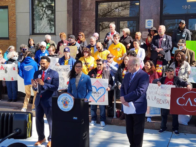Mink is behind a Council podium next to Jawando and Elrich, on the sidewalk in front of the Council building steps, which are filled with community members holding signs supporting the HOME Act. The sun is shining.