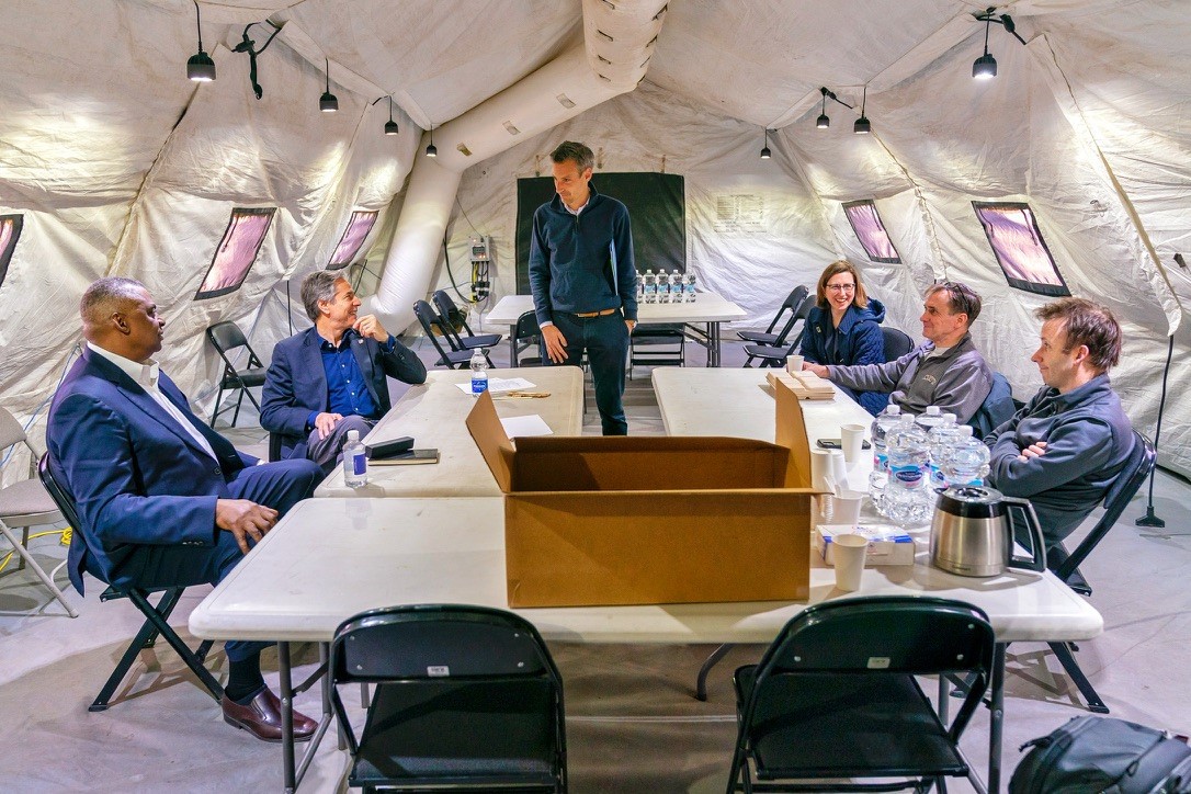 Secretary Blinken is sitting at a table and looking at Spokesperson Price, who is standing. Other U.S. officials are sitting at the table inside a large white tent. 
