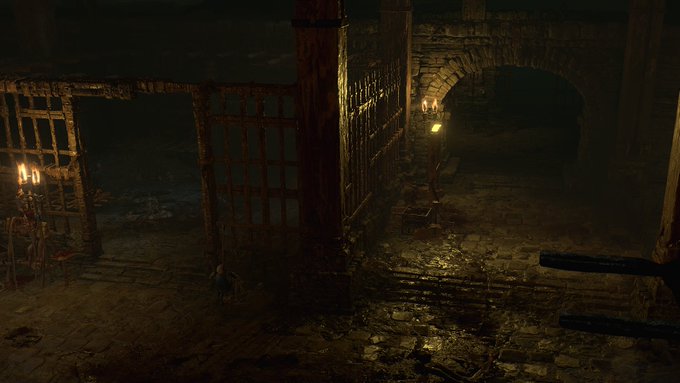 A screenshot of a Diablo 4 dungeon, with a candlestick illuminating what looks like a dank prison, complete with bars and a foreboding  stone entrance.