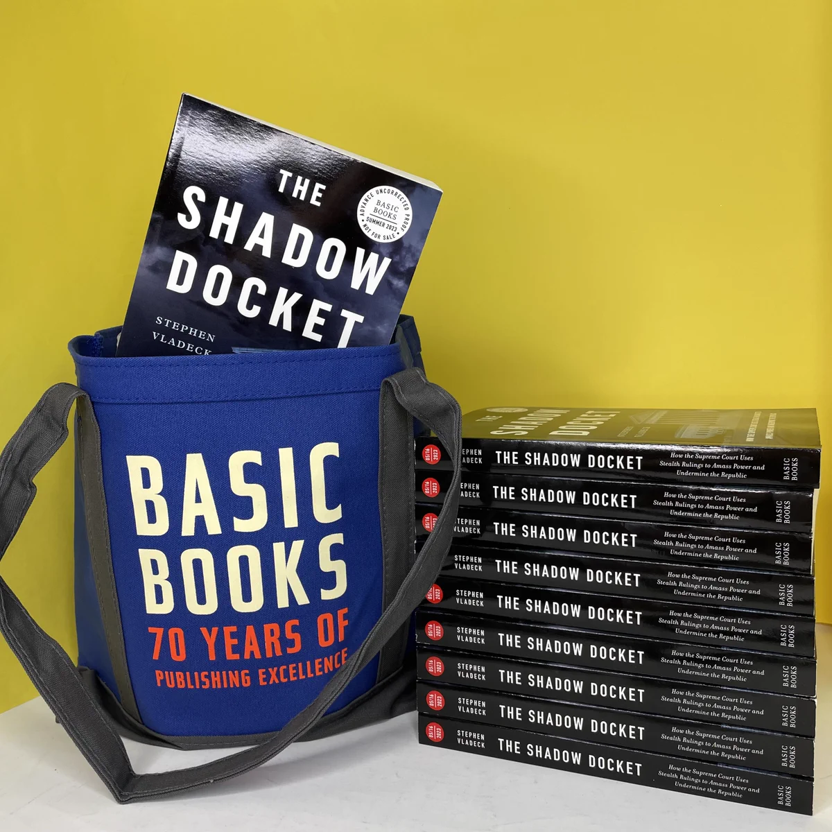 Picture shows a blue tote bag with the words "Basic Books: 70 years of publishing excellence" beside a stack of "The Shadow Docket" book galleys in front of a yellow background.