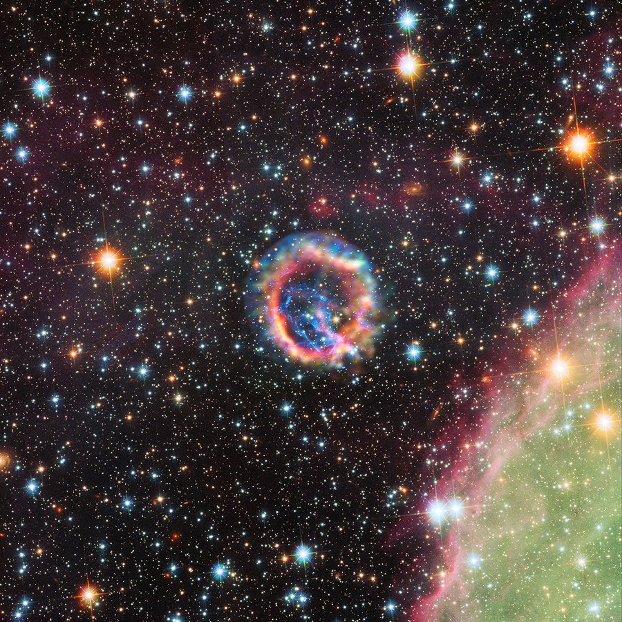 This is a composite image combining X-ray and optical light. Towards the center of the image is a fluffy ring that appears to contain every color in the rainbow. Starting with the innermost portion of the ring, the colors flow outward like mist in shades of red, then orange, then yellow, then green, blue, and purple. In the center of the ring there is a blue structure that resembles lightning. At the lower right of the image, a lime green cloud in the shape of an arc is present. Its inner edge is highlighted in shades of red and pink. Stars of all shapes, sizes, and distances are present throughout the entire image to dazzling effect.