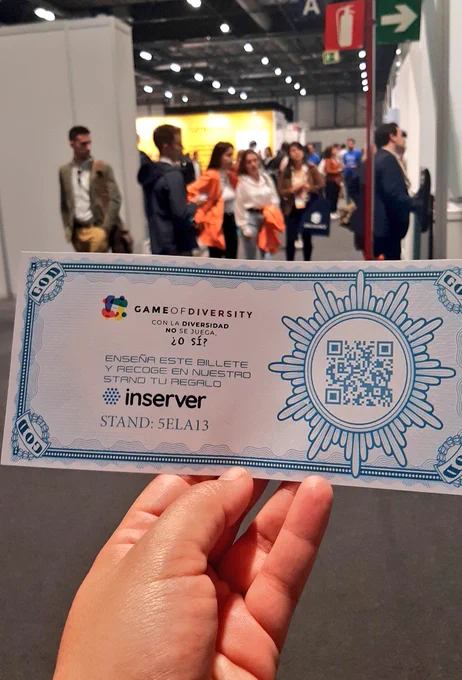 Billete promocional con qr que dirige a la web enlazada en el tuit. Al fondo, marabunta de gente en la feria