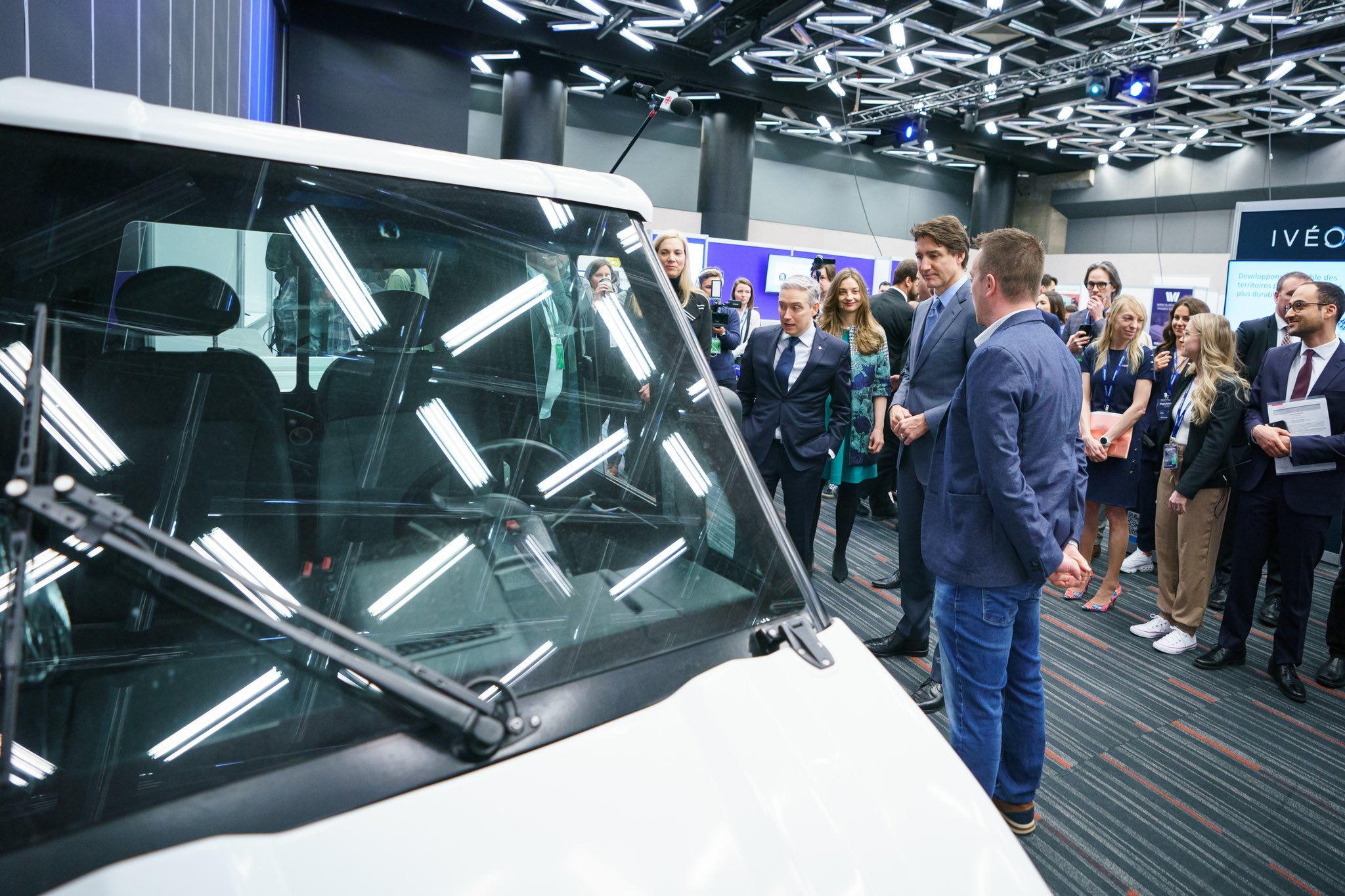 Le premier ministre Justin Trudeau et les ministres François-Philippe Champagne et St-Onge sont debout et discutent avec des gens. Ils regardent un véhicule électrique. D’autres personnes sont debout derrière eux.