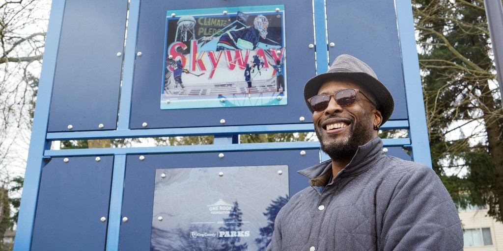 A black man wearing a gray fedora, gray padded felt coat with the collar popped, & brown sunglasses smiles ear to ear in front of a dark blue basketball/hockey goalpost with light blue trim. Behind him is his artwork, showing the word "Skyway" in red superimposed over scenes from the Kraken hockey team playing at Climate Pledge Arena. A shiny plaque also bears the One Roof Foundation & King County Parks logo.