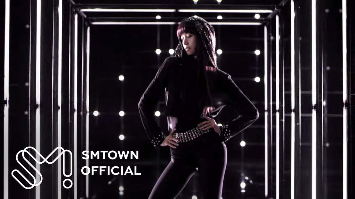 A performer in a black studded hooded outfit and belt poses in a stylized corridor illuminated by bright vertical light tubes. The 'SMTOWN OFFICIAL' logo appears in the bottom left corner.