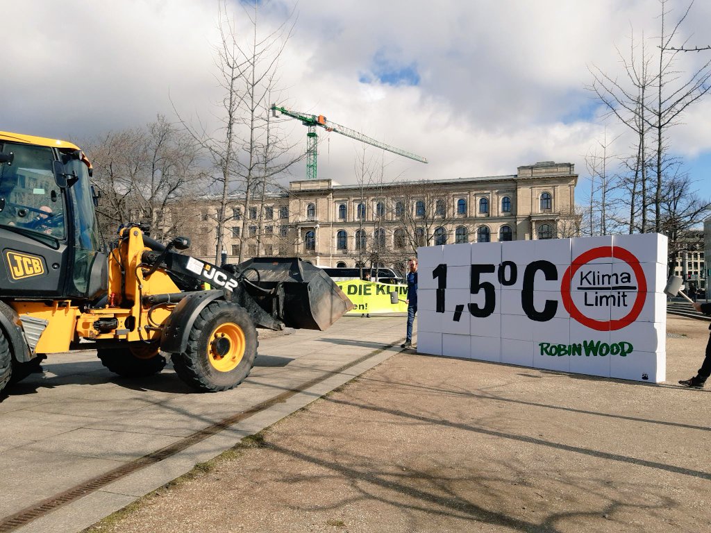 ROBIN WOOD Aktivist*innen protestieren vor dem Bundesverkehrsministerium mit einem Radlader der eine Paketwand mit der Aufschrift "1,5°C Klimalimit" zerstört.