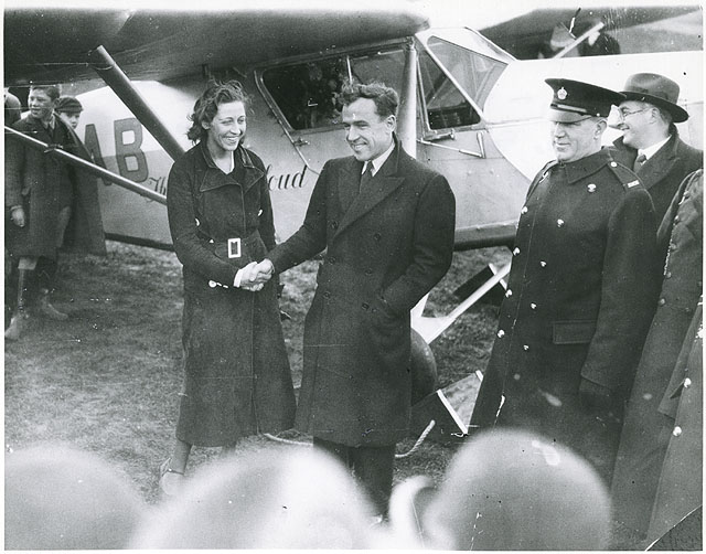 Amy Johnson shakes hands with a man. There is an aircraft behind them.