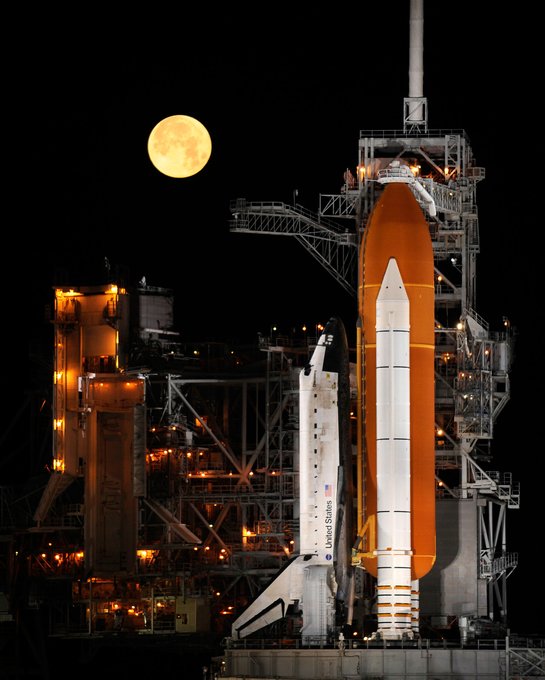 Space Shuttle Discovery attached to the solid rocket boosters and external tank on the launch pad. In the sky above it is a near-full moon.